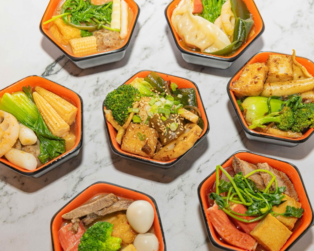 Top-down spread of Fire Wok Hot Pot's seven signature broth bowls — beef bone, tom yum, sukiyaki, tomato, mushroom, pumpkin chicken, and mala xiang guo